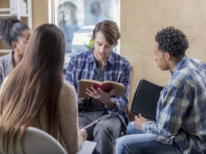 Mid adult Caucasian man concentrates while reading Bible during Bible study. A diverse group of men and women are listening to him read.